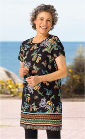 Frau mit kurzen grauen Locken trägt schwarzes Modal-Kleid mit buntem floralen Muster und geometrischem Saum, lächelt, steht vor Meer und Steinpromenade bei hellem Tageslicht
