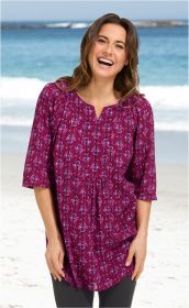 Frau am Strand trägt lächelnd eine violette Viskose-Tunika mit rosa und blauen Mustern, 3/4-Ärmeln und V-Ausschnitt, kombiniert mit dunkler Hose, vor strahlend blauem Meer und weißem Sandstrand.