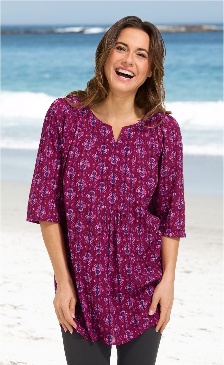Frau am Strand trägt lächelnd eine violette Viskose-Tunika mit rosa und blauen Mustern, 3/4-Ärmeln und V-Ausschnitt, kombiniert mit dunkler Hose, vor strahlend blauem Meer und weißem Sandstrand.