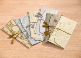 Sechs dekorative Umschläge im Set, verschiedene Beige- und Grautöne mit gold- und silberfarbenen Ornamenten, zum Teil mit Quasten verziert, liegend auf hellem Holztisch
