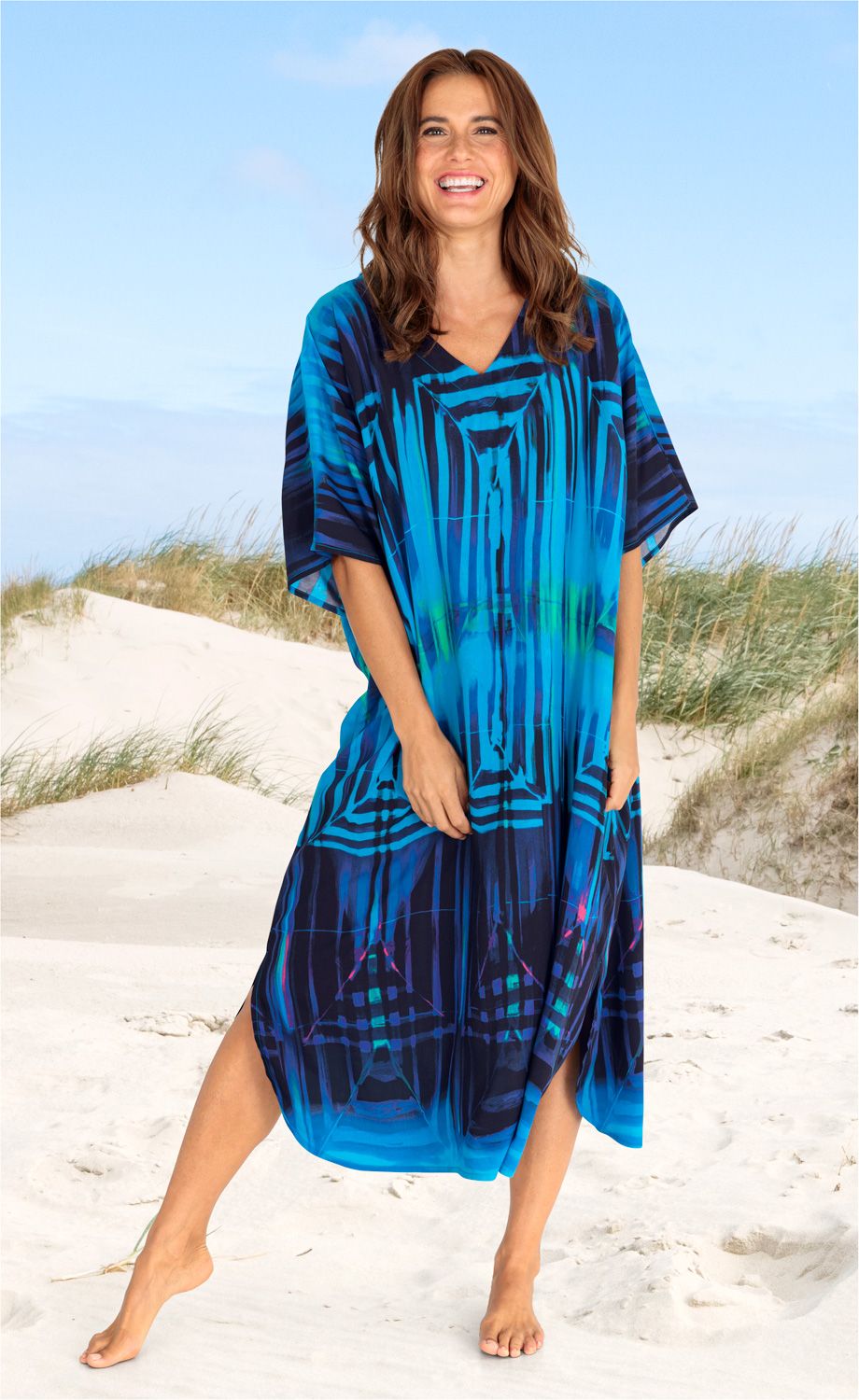 Frau am Strand trägt leichten Viskose-Kaftan in Blau mit abstraktem Muster, knielang, mit V-Ausschnitt, steht barfuß im Sand, lächelt, Hintergrund: Dünen und blauer Himmel