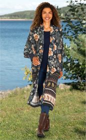 Frau am See trägt lange Viskose-Bluse mit floralem Muster in dunklen Farben, dunkelblaue Jeans, braune Stiefel, lockige Haare, sommerliche Naturkulisse mit Wasser und grüner Wiese im Hintergrund