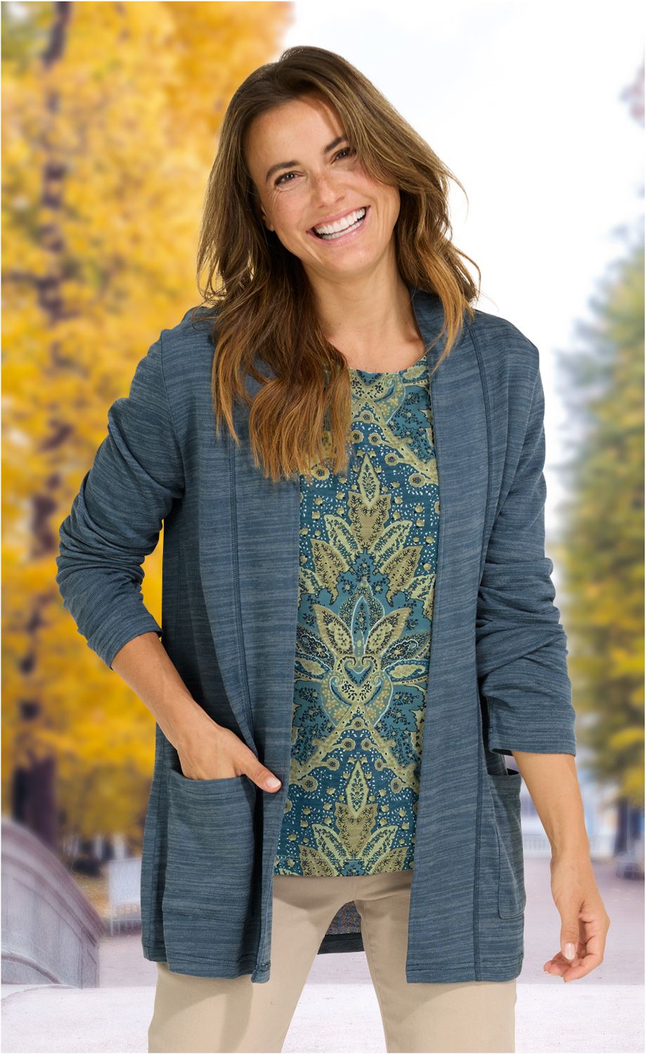 Frau trägt offene blaue Viskose-Mix-Jacke mit langen Ärmeln, gemustertes blau-grünes Shirt, beige Hose, steht lächelnd vor herbstlicher Baumkulisse mit gelben Blättern