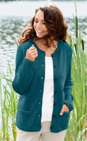 Frau am See trägt dunkeltürkise Alpaka-Strickjacke mit Knöpfen und Taschen, darunter weißes Shirt und helle Hose, lächelt, braune Haare wehen im Wind, grüne Schilfpflanzen im Hintergrund