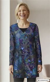 Frau trägt langärmeliges Baumwoll-Mix-Shirt mit V-Ausschnitt, blau-lila Muster mit Punkten und abstrakten floral wirkenden Motiven, lächelnd, steht vor hellem Hintergrund, schwarze Leggings sichtbar