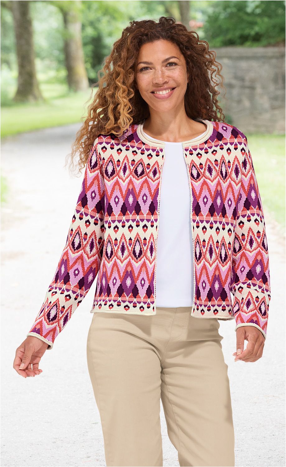 Frau mit lockigen Haaren lächelt, trägt eine bunte Baumwoll-Strickjacke mit geometrischem Muster in Lila, Rosa und Creme, weißes Shirt und beige Hose, steht auf einem hellen Weg mit Bäumen im Hintergrund