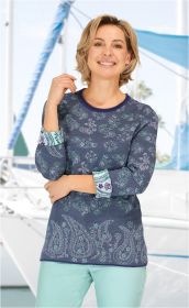 Frau trägt blau gemusterten Baumwoll-Pullover mit langen Ärmeln und floralem Design, hellblau-grüne Manschetten, vor Hintergrund mit Segelbooten und Wasserszene.