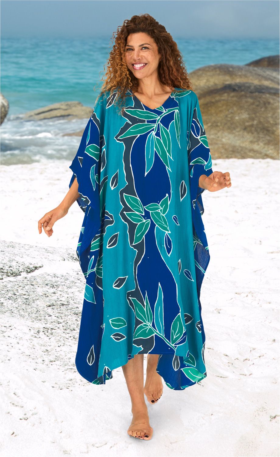 Frau mit lockigem Haar trägt blauen Viskose-Kaftan mit grünem Blattmuster, barfuß am Sandstrand, lächelt, im Hintergrund Meer und Felsen sichtbar