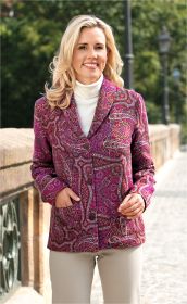 Frau mit blonden Haaren trägt eine bunte Woll-Jacke mit Paisley-Muster in Rot- und Pinktönen, weiße Rollkragenbluse, beige Hose, steht auf gepflastertem Weg neben Steinbalustrade mit grünem Hintergrund.