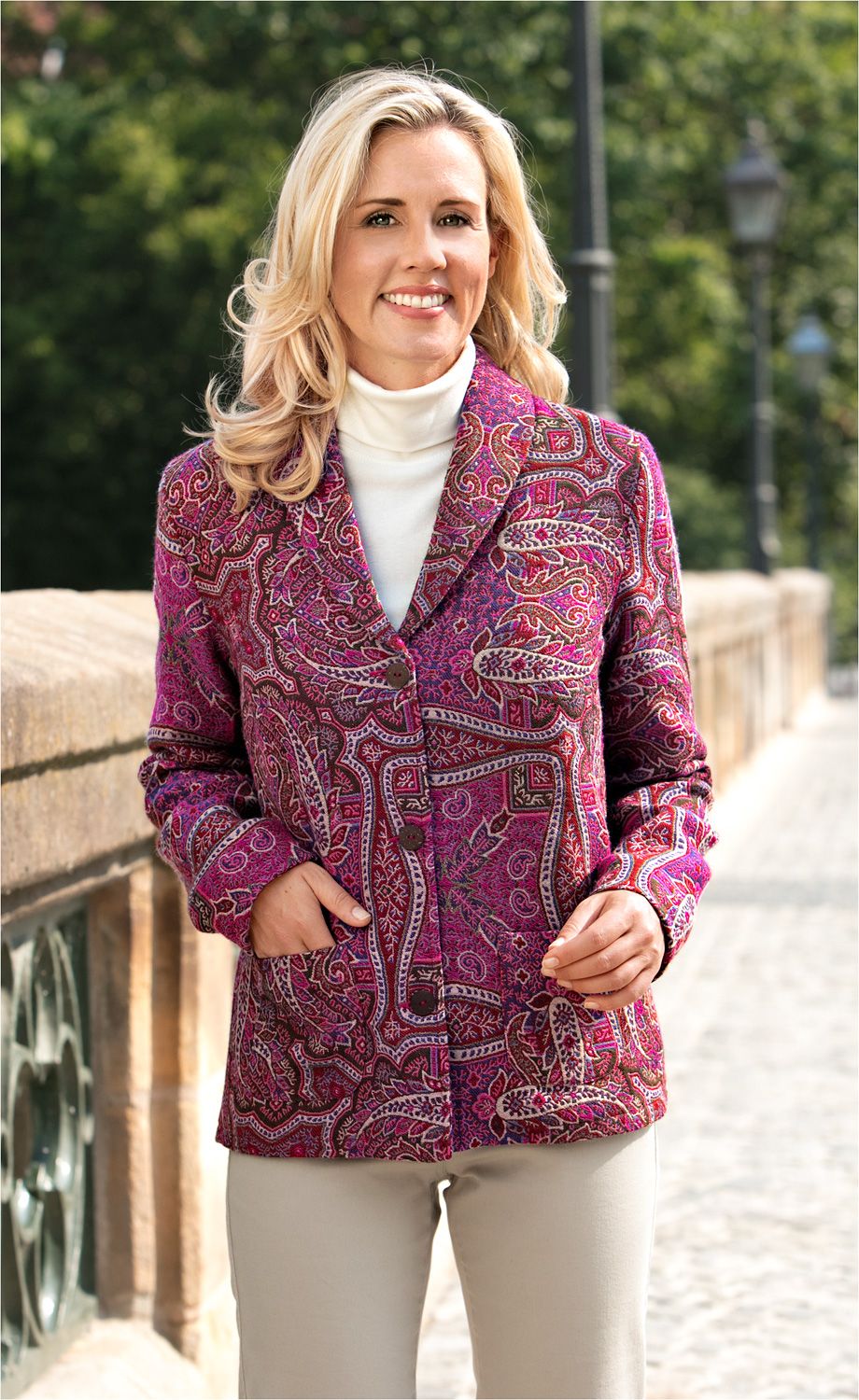 Frau mit blonden Haaren trägt eine bunte Woll-Jacke mit Paisley-Muster in Rot- und Pinktönen, weiße Rollkragenbluse, beige Hose, steht auf gepflastertem Weg neben Steinbalustrade mit grünem Hintergrund.