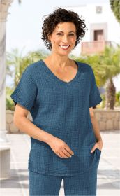 Frau mit kurzen lockigen Haaren trägt blaue Baumwoll-Bluse mit V-Ausschnitt und kurzen Ärmeln, steht vor mediterranem Hintergrund mit Palmen und Mauerwerk, lächelt freundlich, eine Hand in Blusentasche.