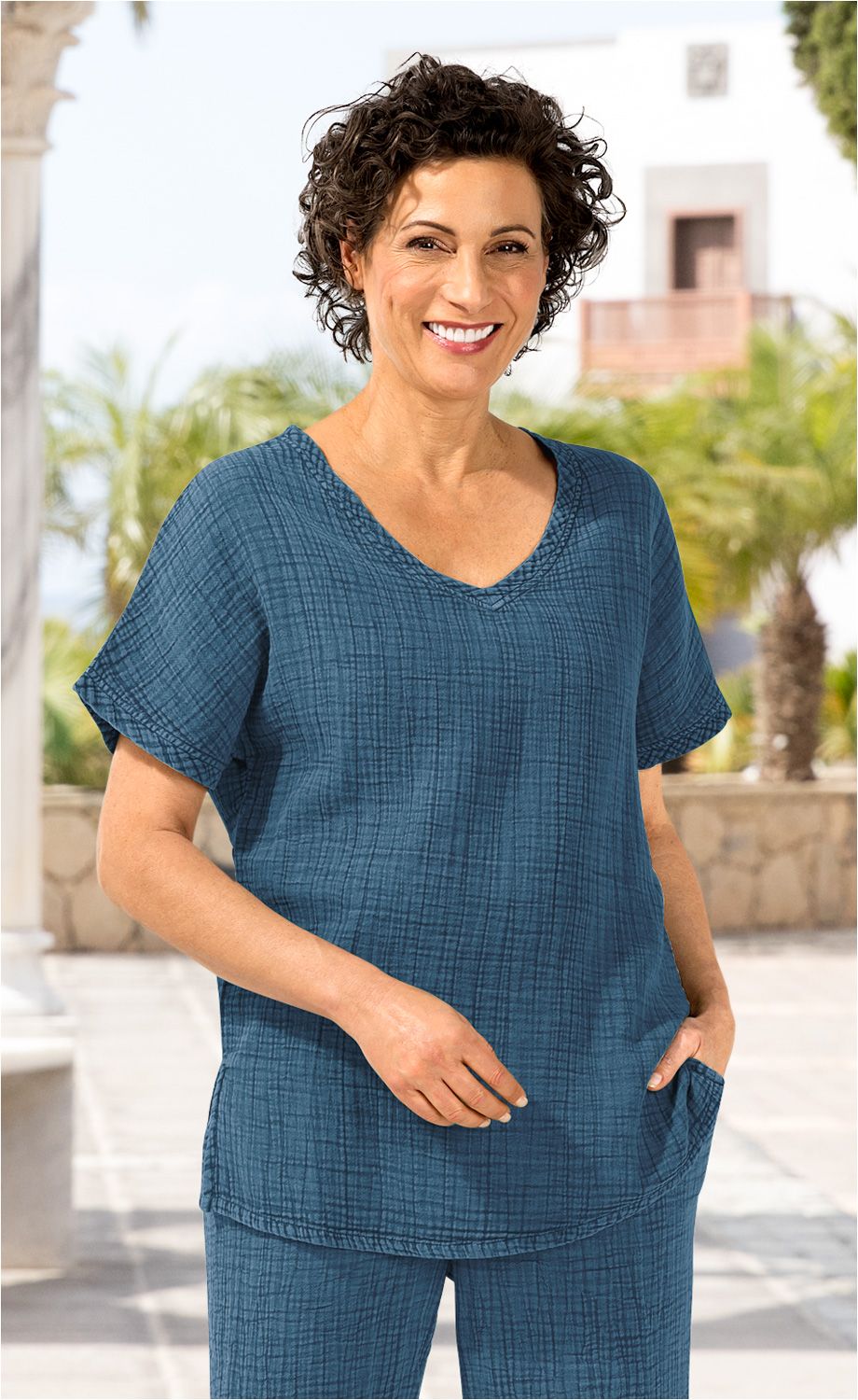 Frau mit kurzen lockigen Haaren trägt blaue Baumwoll-Bluse mit V-Ausschnitt und kurzen Ärmeln, steht vor mediterranem Hintergrund mit Palmen und Mauerwerk, lächelt freundlich, eine Hand in Blusentasche.