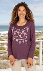 Frau mit langem lockigem Haar trägt ein lila Baumwoll-Shirt mit buntem Laternen- und Blumenmuster, beige Hose, im Hintergrund Meer, Strand und bewölkter Himmel