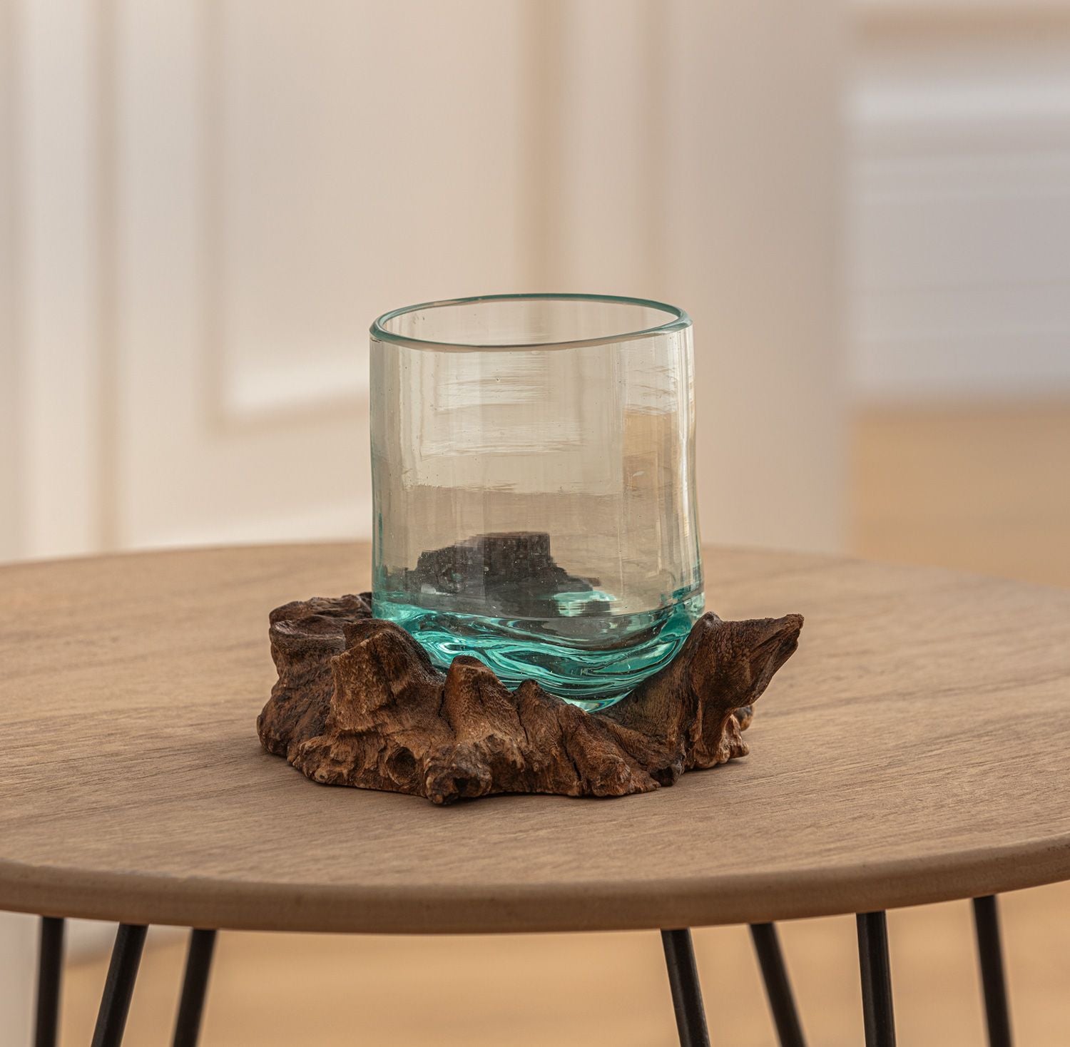 transparentes Windlicht aus Glas mit unregelmäßigem Rand, sitzt auf einem braunen, naturbelassenen Wurzelholzring, steht auf rundem Holztisch mit schwarzen Metallbeinen, heller Hintergrund mit Türen und Wänden unscharf sichtbar