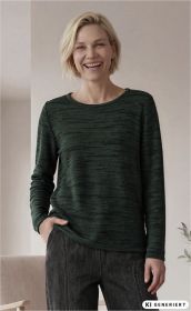 Frau mit kurzem blondem Haar trägt grünen Baumwoll-Pullover mit dezentem Muster, steht entspannt mit einer Hand in der Hosentasche, lächelt, trägt dunkle Jeans, heller neutraler Hintergrund