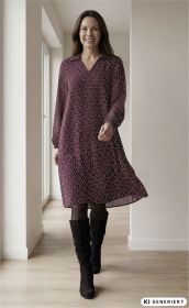 Frau trägt weinrotes, gemustertes Viskose-Kleid mit V-Ausschnitt und Kragen, schwarze Strumpfhose und schwarze kniehohe Stiefel, steht in hellen Raum mit Holzboden und Fenster im Hintergrund, lächelnd
