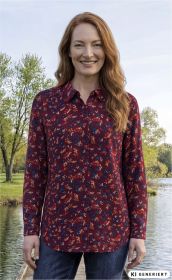 Frau mit roten Haaren trägt rote Viskose-Bluse mit floralem Muster in Blau und Orange, steht lächelnd am Seeufer, bewachsene Bäume im Hintergrund, trägt blaue Jeans