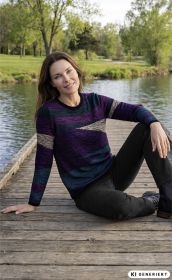 Frau sitzt entspannt auf einem Steg am See, trägt bunt gestreiften Baumwoll-Pullover in Lila, Blau und Beige, schwarze Hose und schwarze Stiefeletten, grüne Bäume und Wasser im Hintergrund, Tageslicht, freundliches Lächeln.