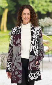 Frau trägt Baumwoll-Mix-Strickjacke mit großem floralen Muster in Schwarz, Grau und Dunkelrot, kombiniert mit weißem Rollkragenpullover und schwarzer Hose, steht vor unscharf grünem Gartenhintergrund