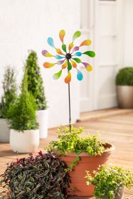 bunter Deko-Gartenstecker mit tropfenförmigen Blättern in Gelb, Grün, Blau und Lila in Terrakotta-Blumentopf auf Holzterrasse, umgeben von grünen und roten Zimmerpflanzen vor weißer Wand und Blumentöpfen
