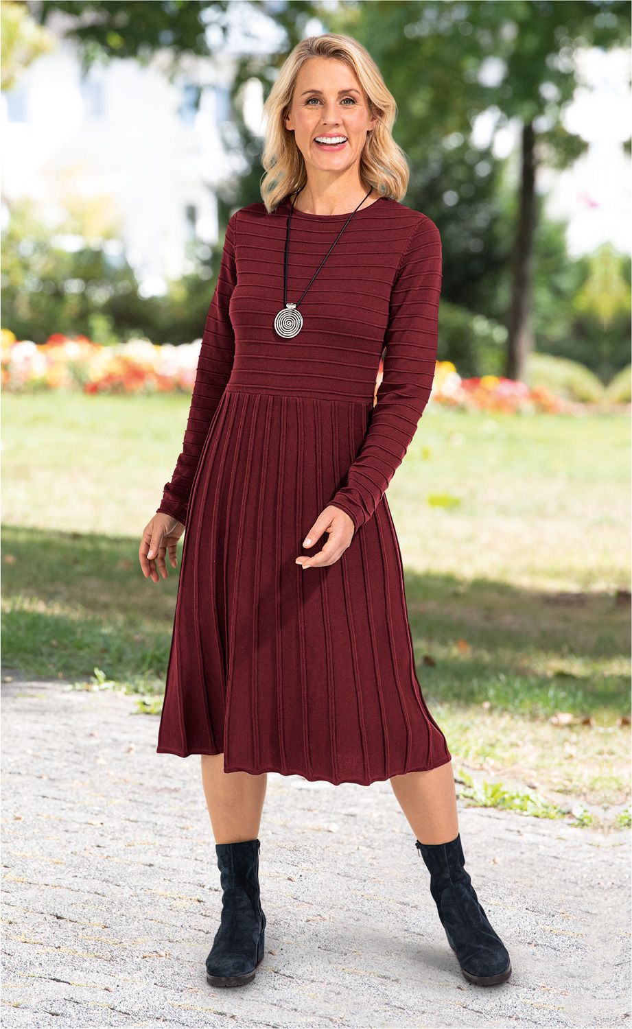 Frau trägt bordeauxrotes, langärmeliges Baumwoll-Kleid mit vertikalen Streifen, knielang, dazu schwarze Stiefeletten und silberne Spiral-Halskette, steht auf gepflastertem Weg, grüner Parkhintergrund, lächelt