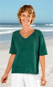 Frau trägt dunkelgrünen Leinen-Mix-Pullover mit V-Ausschnitt und halblangen Ärmeln, weiße Hose, steht lächelnd am Strand mit Meer und blauem Himmel im Hintergrund