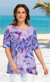 Frau trägt helle Viskose-Tunika mit abstraktem blau-lila Muster, halblange Ärmel, Rundhalsausschnitt mit kleinem Schlitz, weiße Hose, Strand mit Meer und Palme im Hintergrund