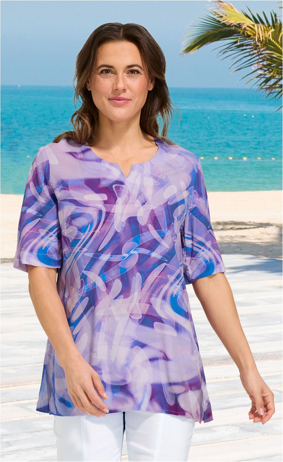 Frau trägt helle Viskose-Tunika mit abstraktem blau-lila Muster, halblange Ärmel, Rundhalsausschnitt mit kleinem Schlitz, weiße Hose, Strand mit Meer und Palme im Hintergrund