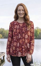 Frau mit roten Haaren trägt eine langärmlige Modal-Bluse in Rot mit großem floralen Muster, steht entspannt vor einem See mit Bäumen im Hintergrund, lächelt, trägt schwarze Hose, natürliche Umgebung, Tageslicht
