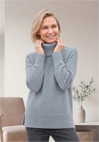 Frau mit kurzem blondem Haar lächelt, trägt grauen Rollkragenpullover und dunkle Hose, steht vor beige Wand mit braunem Sessel, weißer Konsole und Vase mit Zweig, hält Rollkragenpullover an Hals fest