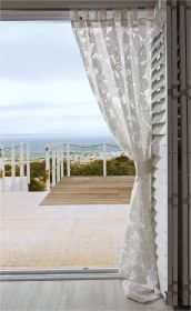 Transparenter Vorhang mit floralem Muster und Schlaufen, leicht zusammengebunden an weißem Klappladen, Blick auf Terrasse mit Holzsteg, Terrasse aus Stein und Meer im Hintergrund bei bewölktem Himmel