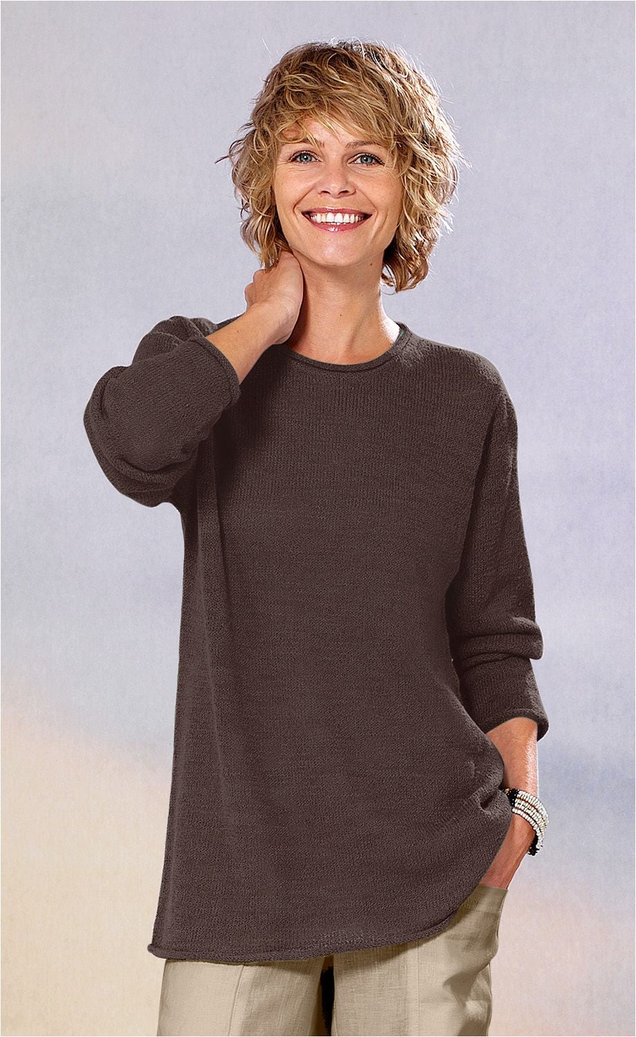 lächelnde frau mit kurzen lockigen blonden haaren, trägt dunkelbraunen pullover und helle hose, steht vor neutralem hintergrund, hand am hals, andere hand in hosentasche, trägt mehrreihiges armband