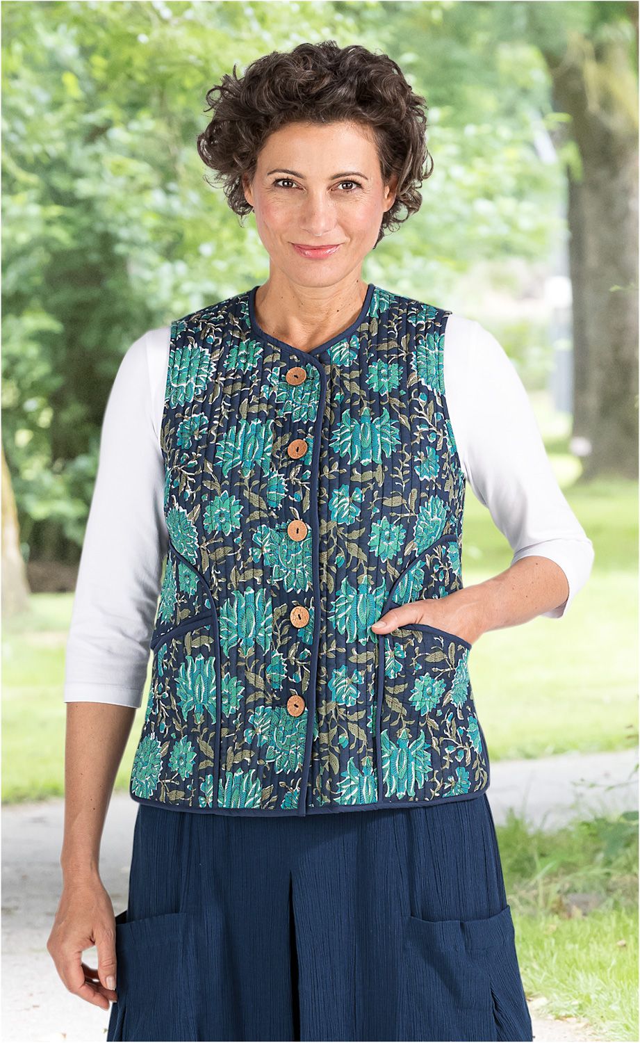 Frau trägt blaue, gesteppte Baumwoll-Weste mit floralem Muster und braunen Knöpfen über weißem 3/4-Arm Shirt, steht im Freien vor grünem, unscharfem Hintergrund
