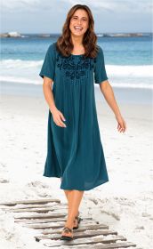 Frau läuft am Strand auf Holzsteg, trägt blaues Viskose-Kleid mit kurzem Arm und floraler Stickerei am Ausschnitt, schwarze Sandalen, lächelt, Hintergrund Meer mit Wellen und bewölktem Himmel