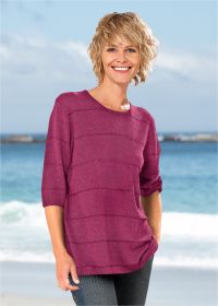 Frau am Strand trägt leuchtend pinken Leinen-Mix-Pullover mit 3/4-Ärmeln, lächelt, kurze blonde Haare, Hintergrund Meer und blauer Himmel
