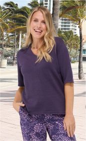 Frau mit blondem, welligem Haar trägt violetten Leinen-Mix-Pullover mit halblangen Ärmeln und gemusterte lila Hose, steht vor Palmen und Hochhäusern, lächelt, eine Hand in Hosentasche, sommerliches Stadtbild im Hintergrund