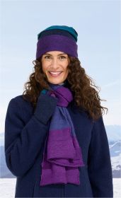 Frau mit lockigen braunen Haaren trägt blauen Mantel, bunt gestreiften lila-blauen Alpaka-Schal und passende Mütze, lächelt vor winterlicher Berglandschaft mit Schnee und klarem Himmel