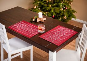 Dunkler Holztisch mit weißem Holzgestell, zwei weiße Stühle, rotes Baumwoll-Tischset 2er-Set mit weißem Weihnachtsschriftmuster, auf dem Tisch Glas mit Tee, Kerze auf Holzstern, im Hintergrund geschmückter Weihnachtsbaum