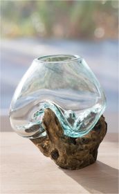 Transparente Glasvase mit geschwungener Form, die auf einem naturbelassenen, knorrigen Holzsockel sitzt, auf hellem Untergrund vor unscharfem, mehrfarbigem Hintergrund.
