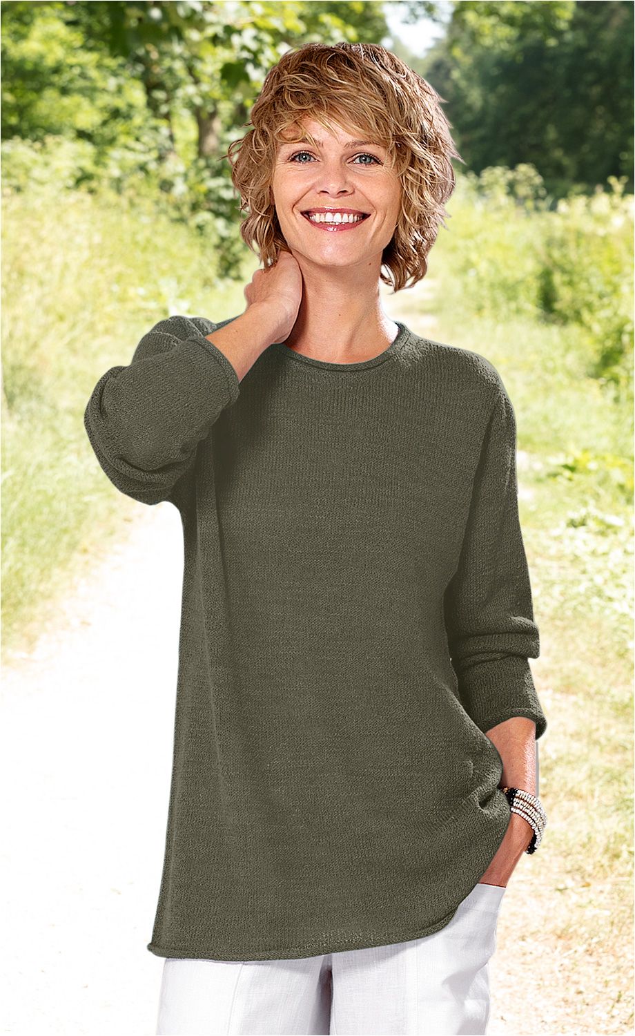 Frau mit kurzen blonden Haaren trägt olivgrünen Baumwoll-Pullover mit langen Ärmeln und weißer Hose, steht lächelnd auf einem Weg im Grünen, eine Hand am Nacken, andere in der Hosentasche, trägt mehrreihiges Armband am Handgelenk
