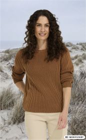 Frau mit langen lockigen braunen Haaren trägt braunen gestreiften Pullover und helle Hose, steht am Strand mit Sanddünen und grauem Himmel im Hintergrund, lächelt freundlich