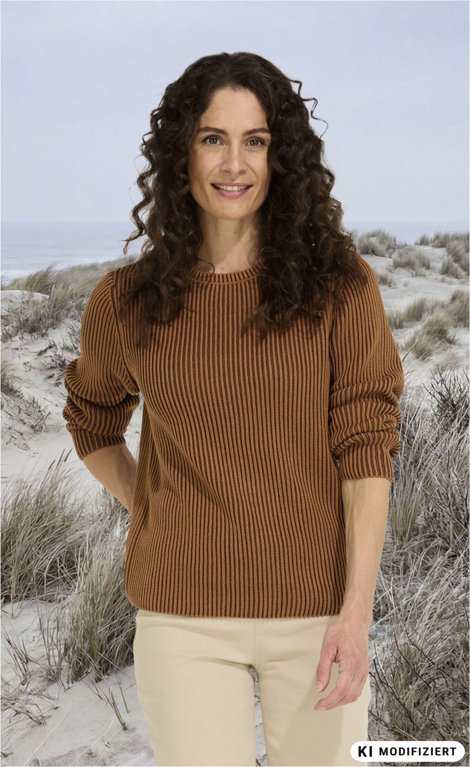 Frau mit langen lockigen braunen Haaren trägt braunen gestreiften Pullover und helle Hose, steht am Strand mit Sanddünen und grauem Himmel im Hintergrund, lächelt freundlich