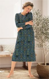 Frau trägt blau-grünes Baumwoll-Hauskleid mit floralem Muster und langen Ärmeln, steht barfuß in hellem Wohnzimmer mit Holzboden, Steppteppich, Bank mit Kissen und großer Pflanze in Korb