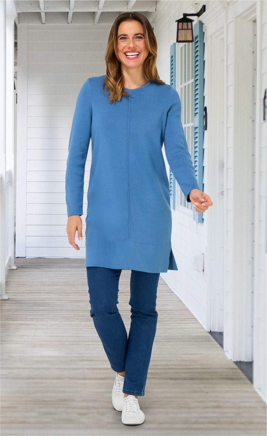 Frau in locker sitzendem, langärmligem blauem Baumwoll-Mix-Kleid, dunkelblauer Jeanshose und weißen Sneakers, lachend, auf hellem Verandaboden mit weißen Wänden und blauen Fensterläden gehend