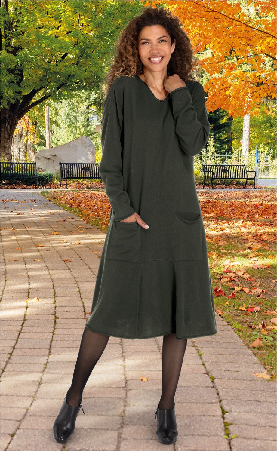 lächelnde frau mit lockigem haar trägt dunkelgrünes langärmeliges kleid mit taschen, schwarze strumpfhose und schwarze schuhe, steht auf gepflastertem weg, herbstliche parklandschaft mit buntem laub und bäumen im hintergrund