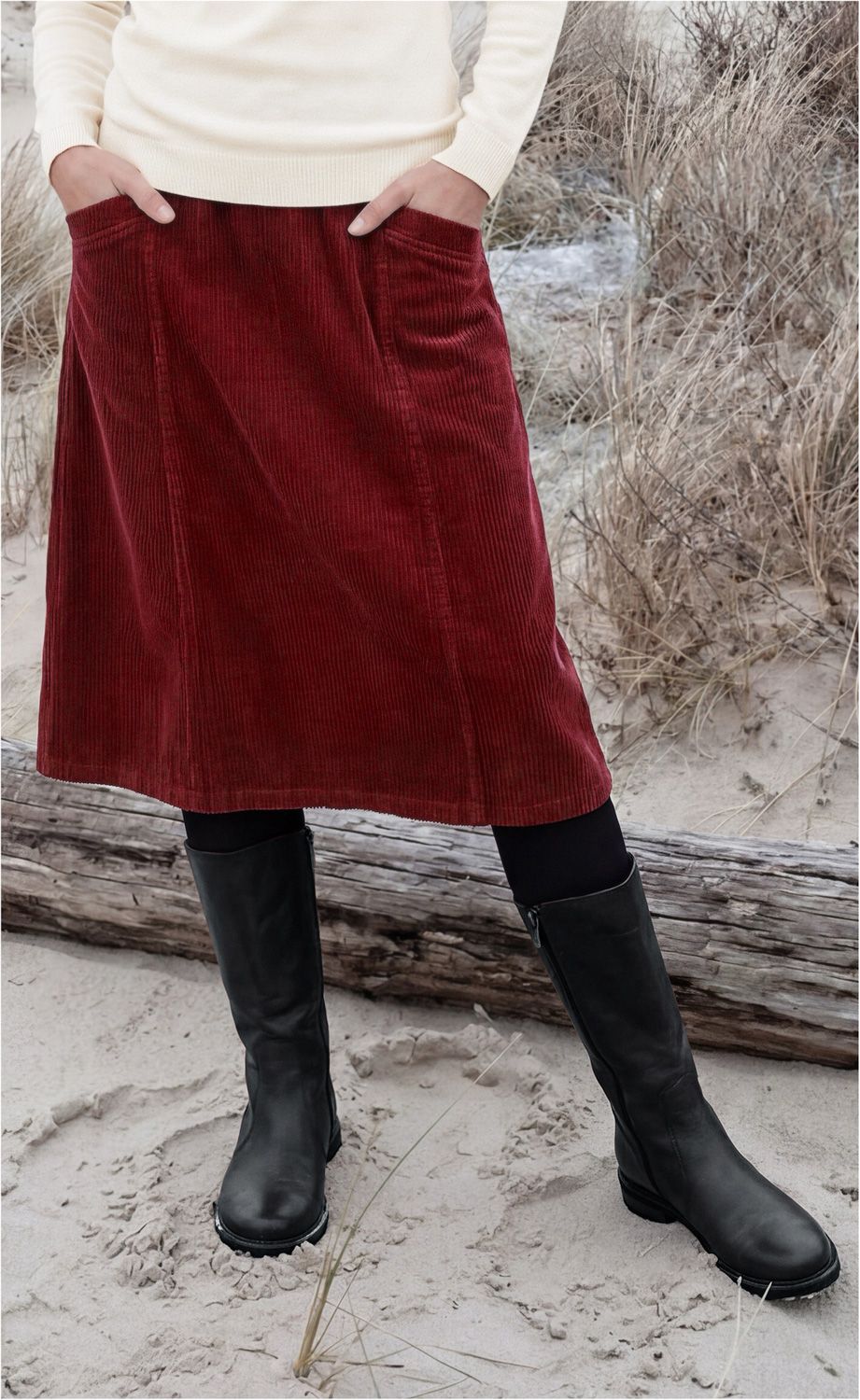 Person steht im Sand, trägt roten Cordrock mit Taschen, schwarzen Strumpfhosen, hohe schwarze Lederstiefel und cremefarbenen Pullover, Hintergrund mit trockenem Gras und Treibholz