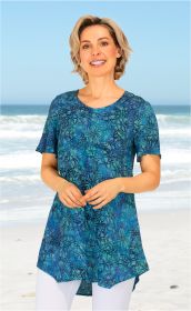 Frau an Strand trägt blau-grüne Viskose-Bluse mit floralem Muster, kurzärmelig, langer, asymmetrischer Schnitt, kombiniert mit weißen Hosen, lächelt, blonder kurzer Haarstil, klarer Himmel und Meereswellen im Hintergrund