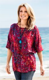 Frau am Strand trägt bunte Viskose-Bluse mit halblangen Ärmeln und Kordelzug in Taille, blau-rot gemustert, dazu lange Halskette mit großen blauen Anhängern, dunkle Jeans