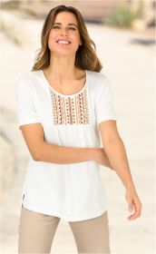 Frau trägt weißes Baumwoll-Shirt mit kurzen Ärmeln und buntem, geometrischem Stickmuster auf der Brust, kombiniert mit beiger Hose, lächelnd vor hellem, unscharfem Hintergrund
