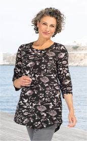 Frau mit kurzen lockigen Haaren trägt schwarze Baumwoll-Tunika mit grauem Blumenmuster und seitlichen Knöpfen, steht am Wasser auf Holzsteg, lächelnd, helle Hintergrundbebauung, hellgraue Hose